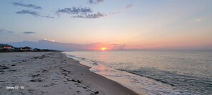 Une plage où l’on voit des empreintes de pas menant à l’eau, un coucher de soleil en arrière-plan et des maisons au loin.