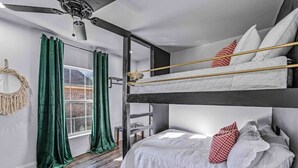 Une chambre avec un lit superposé, un ventilateur de plafond, des rideaux verts et une vue sur un bâtiment en briques à l’extérieur.