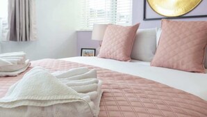 Ein ordentlich bezogenes Bett mit rosa und weißer Bettwäsche, ein Kunstwerk in goldener Rahmung an der Wand und ein Fenster mit Jalousien.