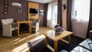 Un salon moderne avec une table à manger en bois, deux fauteuils, un canapé, un téléviseur fixé sur un panneau en bois et une grande fenêtre avec des rideaux.