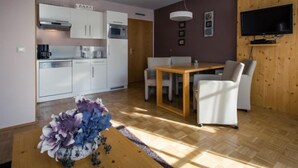 Une cuisine moderne avec des meubles blancs, un four à micro-ondes et une machine à café. On y trouve un coin repas avec une table en bois et des chaises blanches. Un téléviseur est fixé au mur, et un vase rempli de fleurs est posé sur la table.