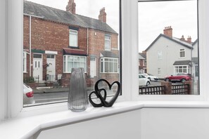 A window with a view of a residential street, a clear sky, and a white wall with a black heart-shaped object. - Stylish 3 Bed House With TVs in Each Bedroom (Darlington)