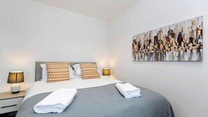 A hotel room with a bed, two bedside lamps, a painting on the wall, and a neatly folded towel on the bed. - Stylish 3 Bed House With TVs in Each Bedroom (Darlington)