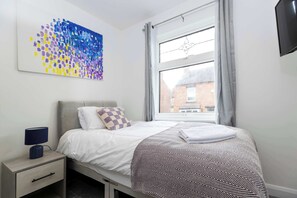 A bedroom with a bed, a nightstand, a lamp, a window with curtains, and a wall art piece.