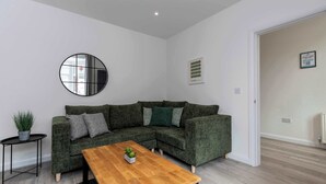 A modern living room with a sectional sofa, a round mirror, a small table with a plant, and a framed picture on the wall. - Stylish 3 Bed House With TVs in Each Bedroom (Darlington)