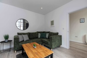 Ein modernes Wohnzimmer mit einer Eckcouch, einem runden Spiegel, einem kleinen Tisch mit einer Pflanze und einem gerahmten Bild an der Wand.