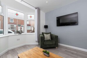 A modern living room with a grey wall, a flat-screen TV mounted above a green sofa, a wooden coffee table with a plant, and a window with decorative glass panes.