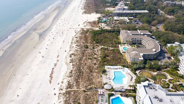 An aerial view of a coastal resort with a long sandy beach, a large pool area, and multiple buildings with balconies.
