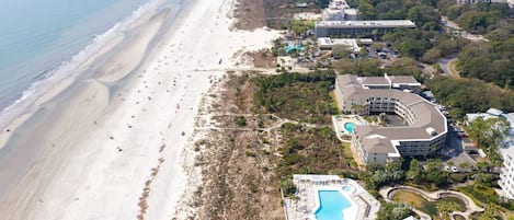 An aerial view of a coastal resort with a long sandy beach, a large pool area, and multiple buildings with balconies.