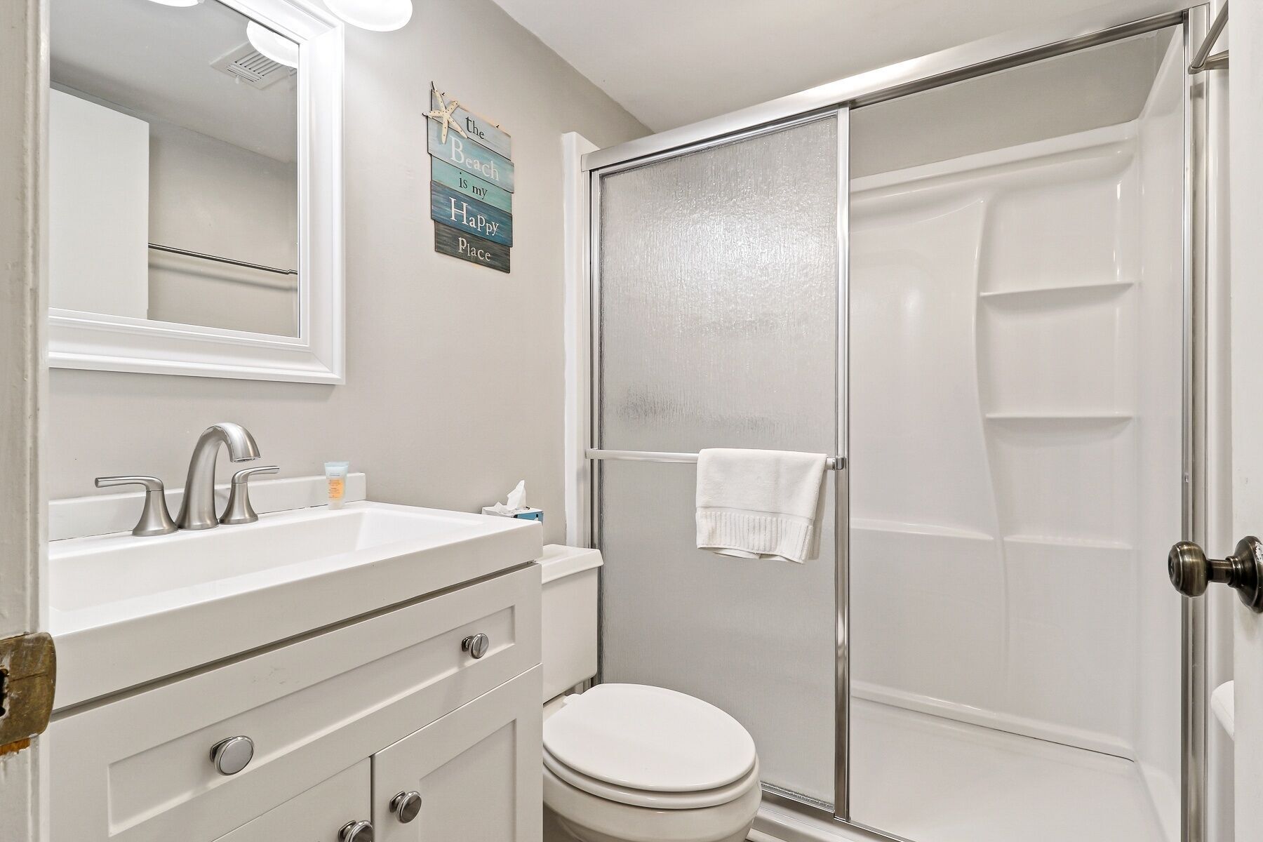 A bathroom with a white sink, mirror, and toilet. There is a shower with a glass door and a towel hanging on the door.