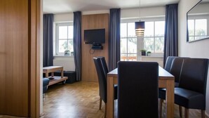 Uma sala de jantar moderna com uma mesa de madeira e cadeiras pretas, uma televisão na parede e janelas amplas com cortinas.