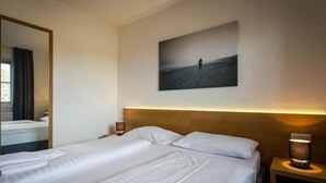 Quarto de hotel com uma cama grande, duas mesas de cabeceira, um espelho e um quadro de uma pessoa numa praia.
