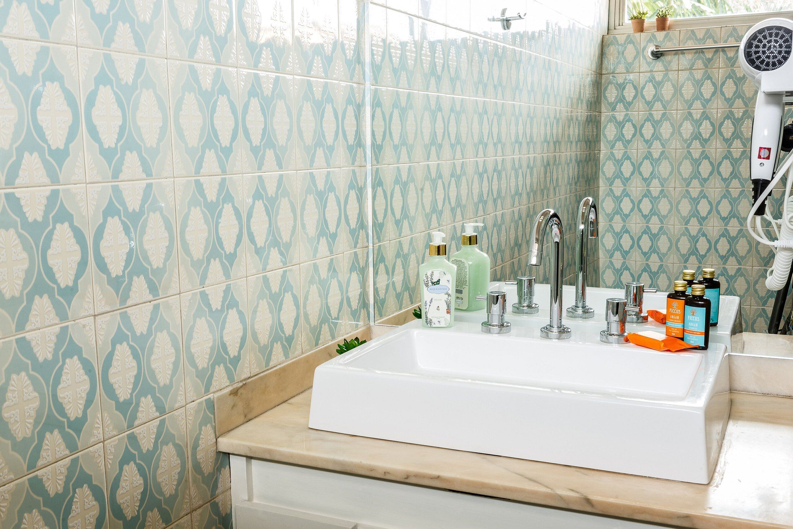 Bagno con lavandino bianco, due rubinetti e soffione doccia a mano. Sul ripiano del lavandino ci sono articoli da toilette.