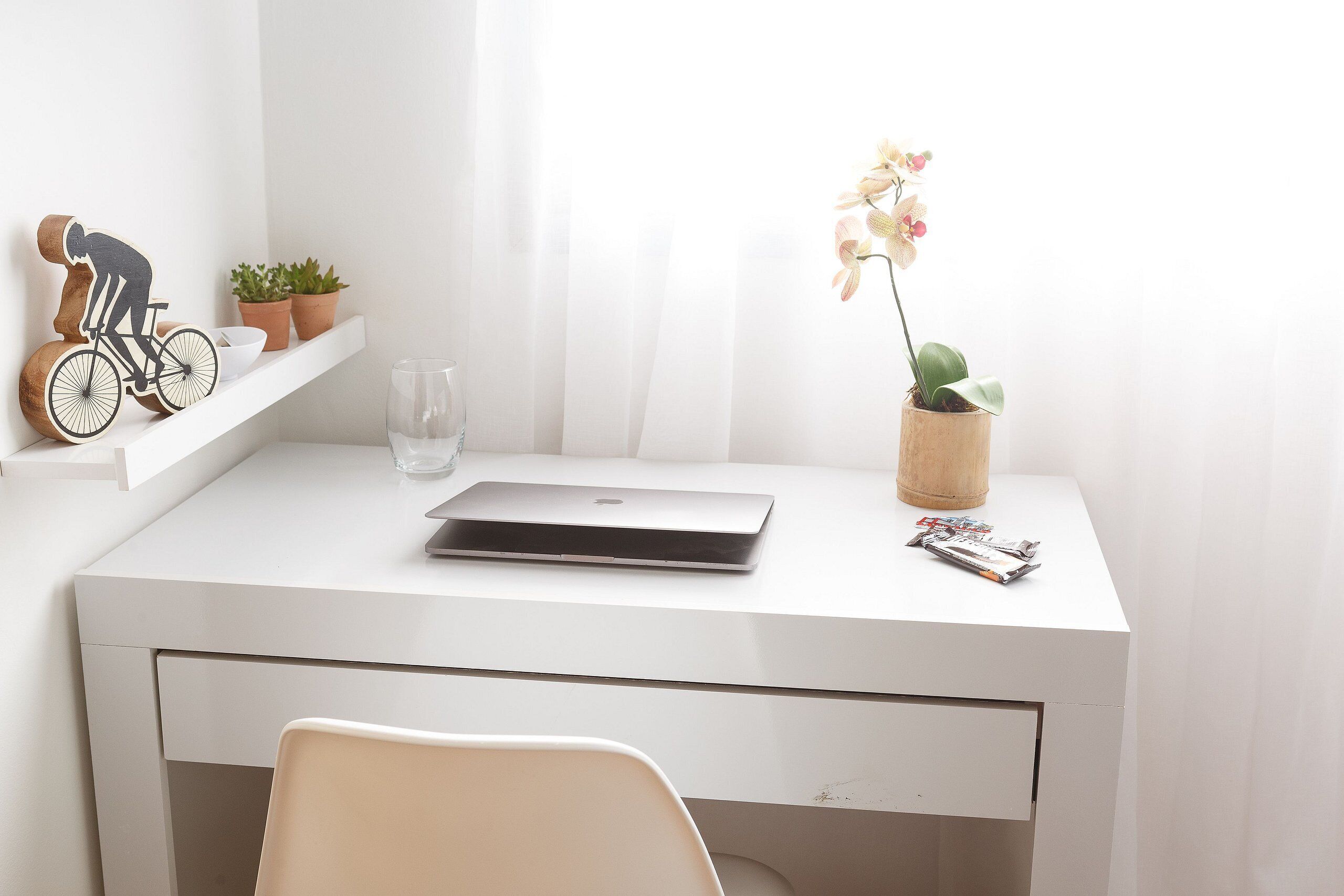 Uma secretária branca com um laptop, uma orquídea em vaso e uma escultura de madeira de um ciclista.