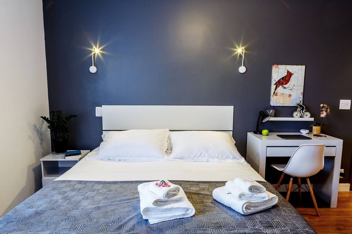 A neatly made bed with white linens, a desk with a chair, a wall-mounted light fixture, and a framed picture.