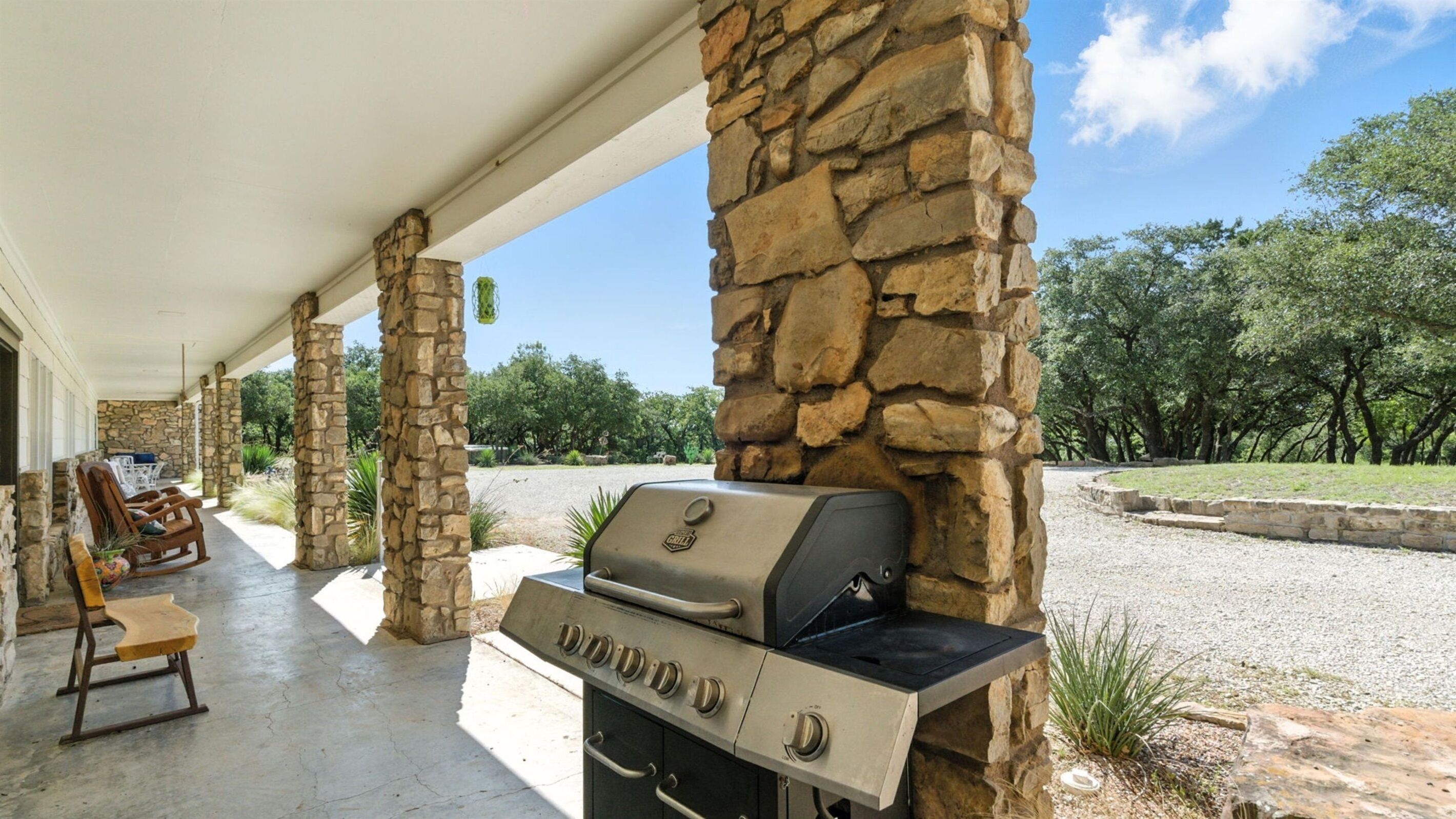 Un patio cubierto con pilares de piedra, una parrilla, mobiliario exterior y vista a árboles.