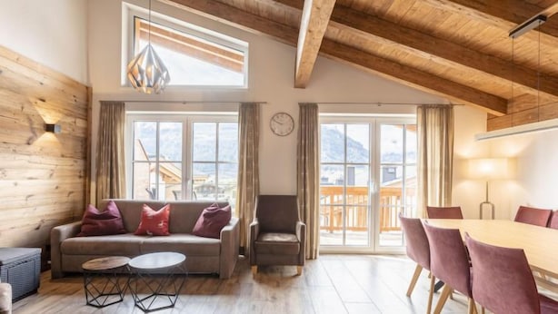 Una sala de estar acogedora con un sofá, un sillón y una mesa de comedor. Amplias ventanas ofrecen vistas al exterior.