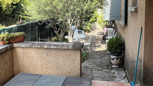 Eine überdachte Terrasse mit Tisch und Stühlen, einem Blumentopf und Blick auf einen Garten mit Bäumen sowie ein Gebäude im Hintergrund.