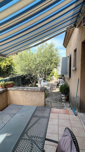A covered patio with a table and chairs, a potted plant, and a view of a garden with trees and a building in the background.