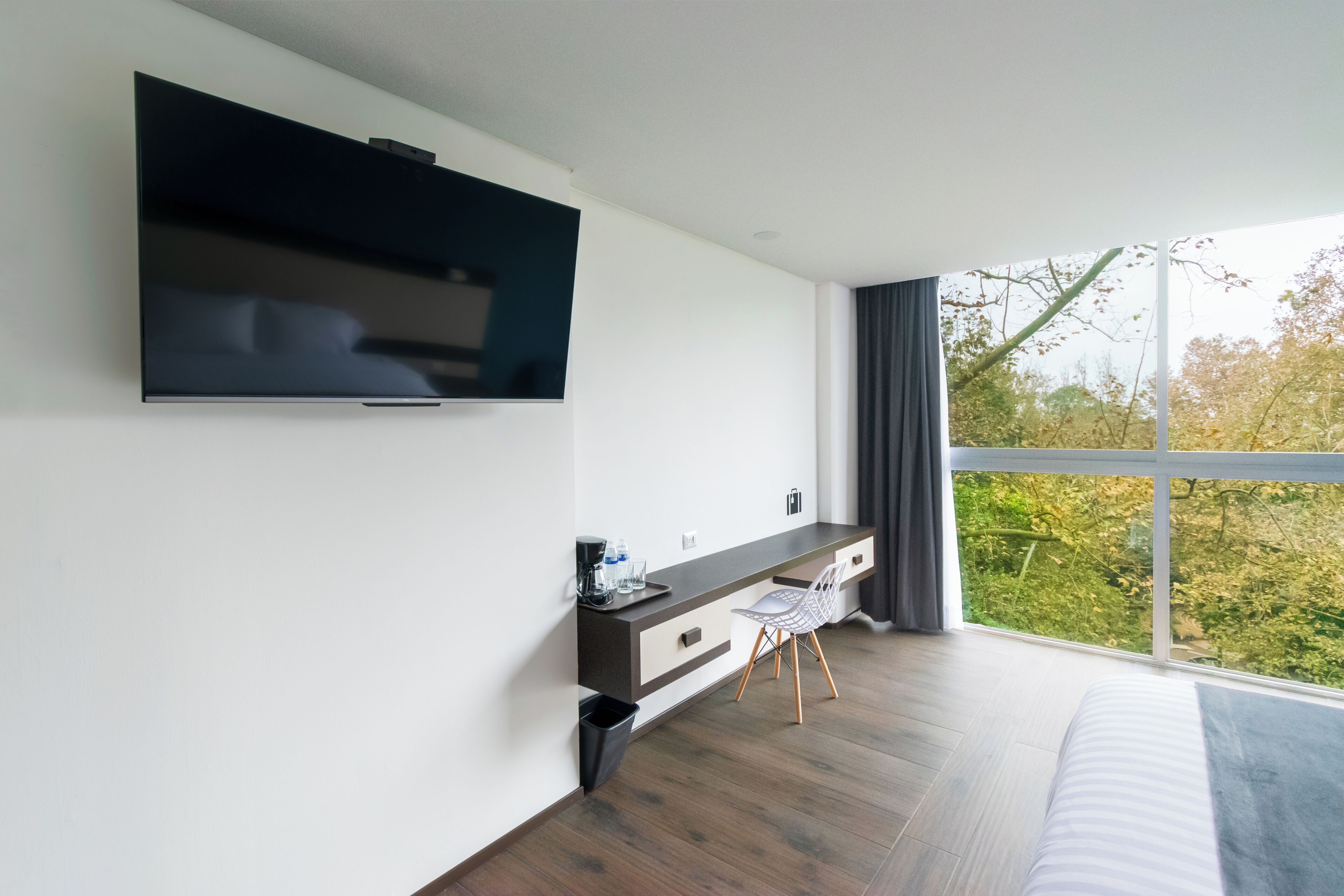 A modern bedroom with a large window, a flat-screen TV mounted on the wall, a bedside table with a lamp, and a bed with a headboard.