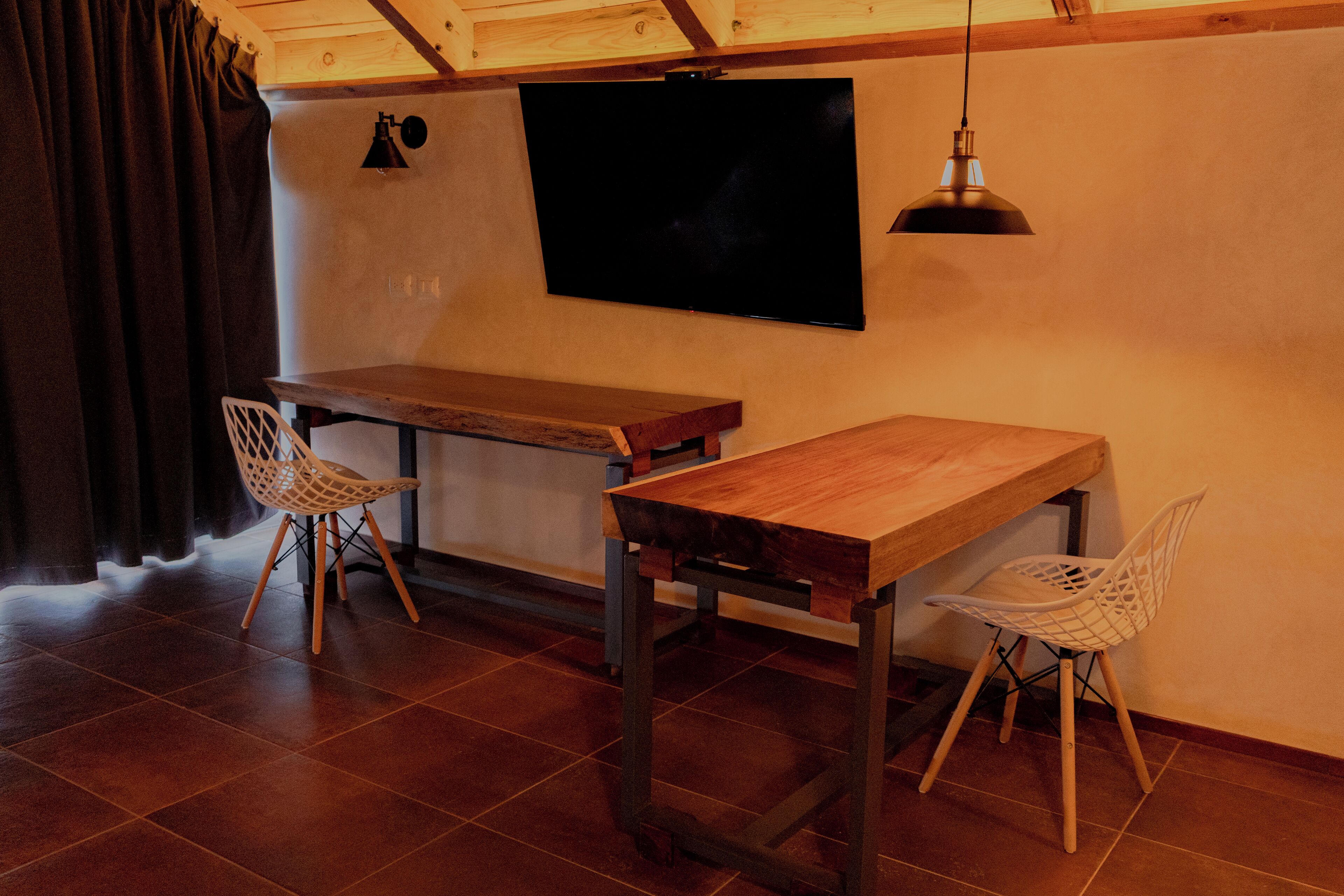 A room with a wooden table and two white chairs, a TV mounted on the wall, and a hanging light fixture.