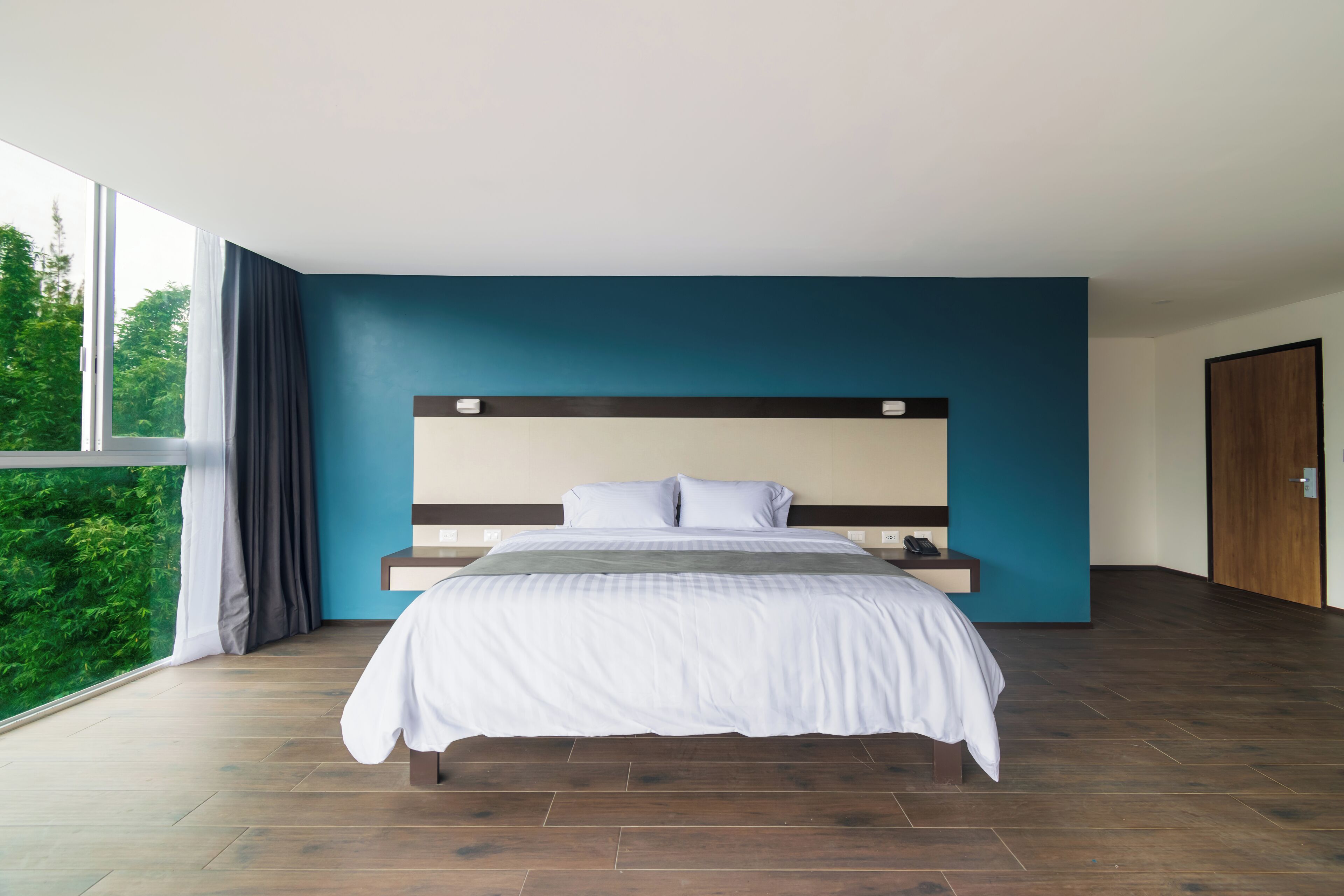 A modern bedroom with a large bed, a view of greenery through a window, and a wooden floor.