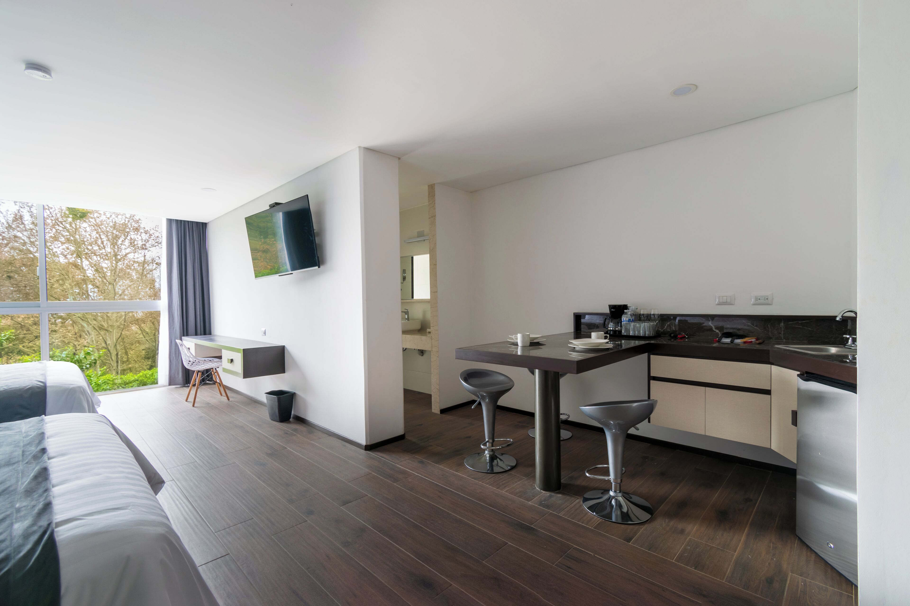A modern hotel room with a large bed, a desk, a kitchen area with bar stools, and a TV mounted on the wall.