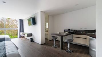 A modern hotel room with a large bed, a desk, a kitchen area with bar stools, and a TV mounted on the wall.