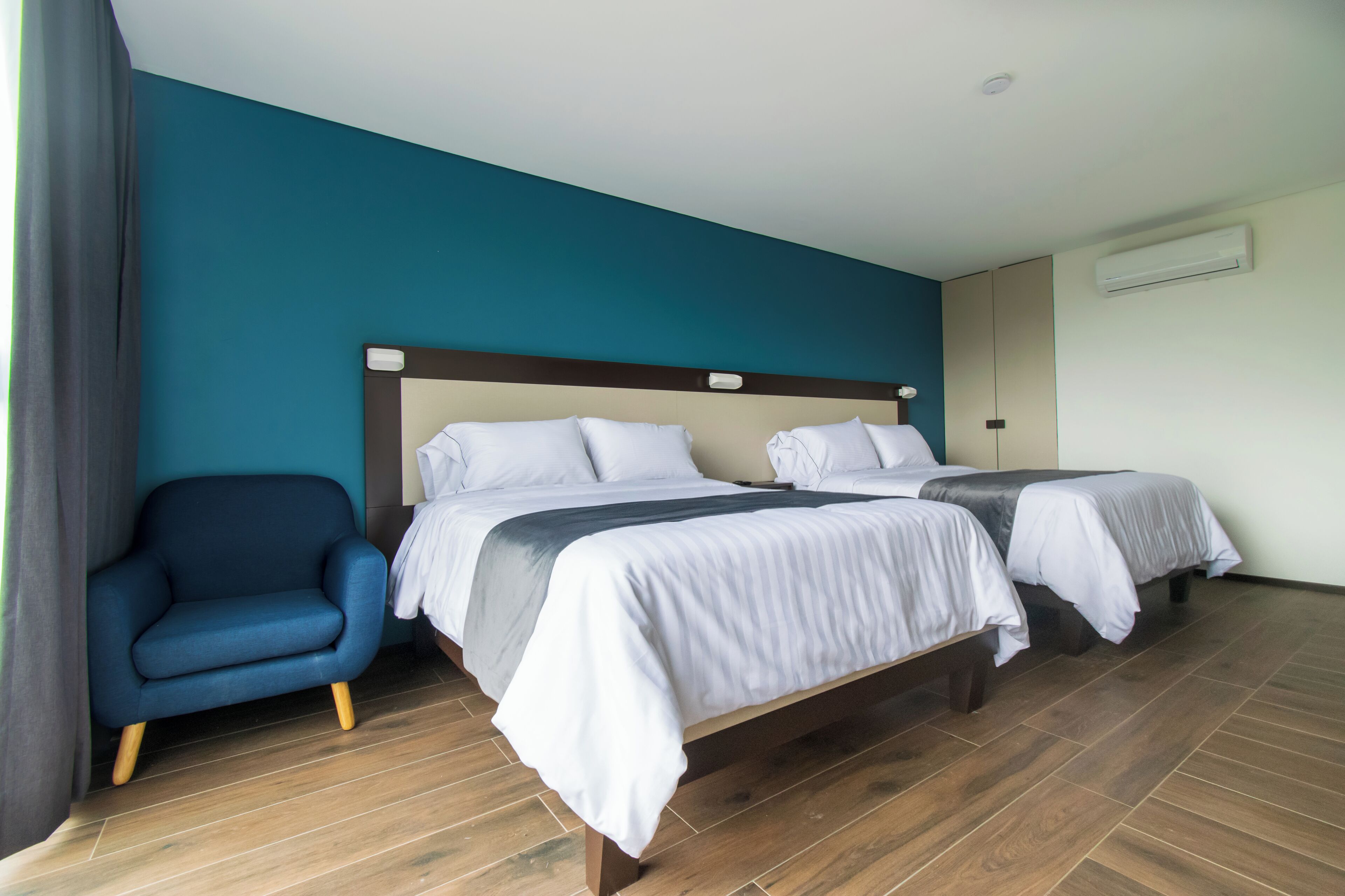 A hotel room with two beds, a blue armchair, and a wooden floor.