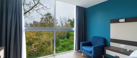 Una habitación de hotel moderna con un ventanal grande, un sillón azul, una cama y una mesita de noche.