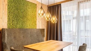 Un espace repas moderne avec une table en bois et deux chaises rembourrées, un mur texturé de couleur verte et une grande fenêtre ornée de rideaux légers.