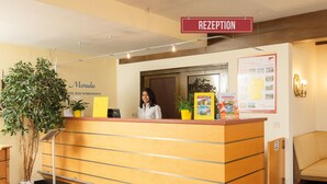 Un mostrador de recepción con un cartel que dice "RECEPTION". A la izquierda del mostrador hay una planta, y detrás, en la pared, un tablón de anuncios con varios elementos y un mapa.