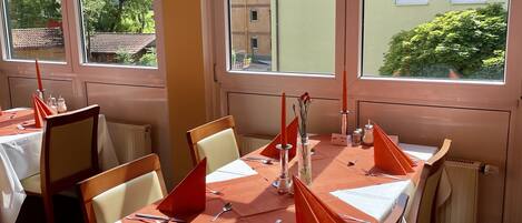 A dining area with tables set for four, large windows, and a view of buildings and trees outside.