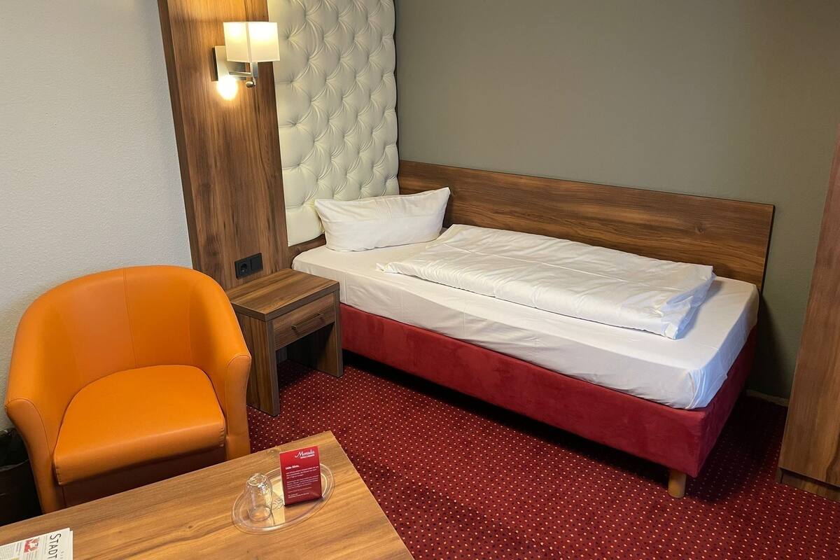 Una habitación de hotel con una cama, una mesita de noche de madera, una alfombra roja, una silla de madera y una mesa con un menú.