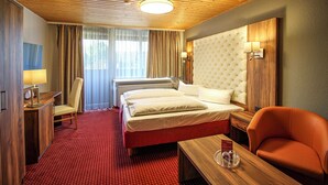 Una habitación de hotel con techo de madera, una cama grande con ropa blanca, un escritorio de madera con lámpara, alfombra roja y un sillón rojo.