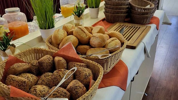 Una mesa de buffet con panes y pasteles varios, un dispensador de zumo y plantas decorativas.