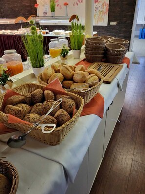 Una mesa de buffet con panes y pasteles varios, un dispensador de zumo y plantas decorativas.