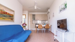 Een moderne woonkamer met een blauwe bank, een eethoek met twee stoelen, een keuken met lichte kastjes, en een tv op een witte dressoir.