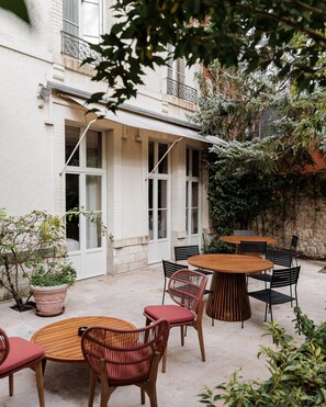 An outdoor patio area with round wooden tables and chairs, a potted plant, and a building with large windows and a balcony. - Frenchdoor Orléans Arago (Orléans)
