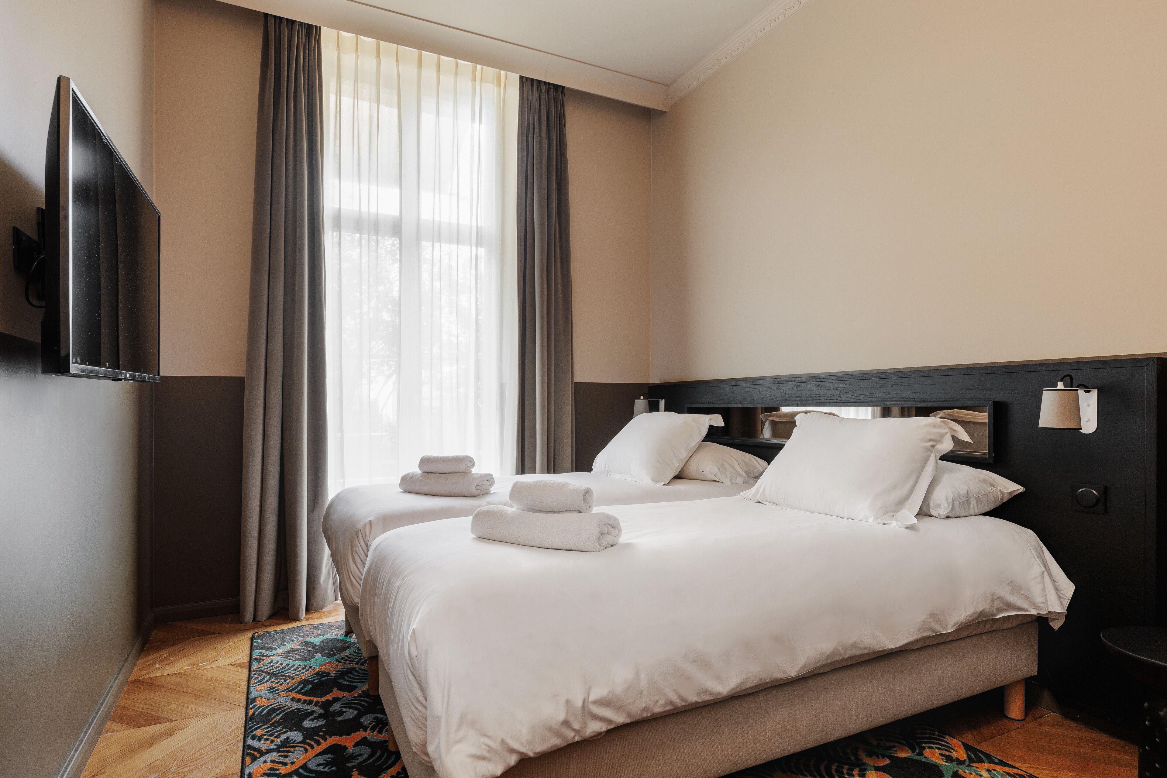 A hotel room with two beds, a flat-screen TV mounted on the wall, and a large window with curtains.