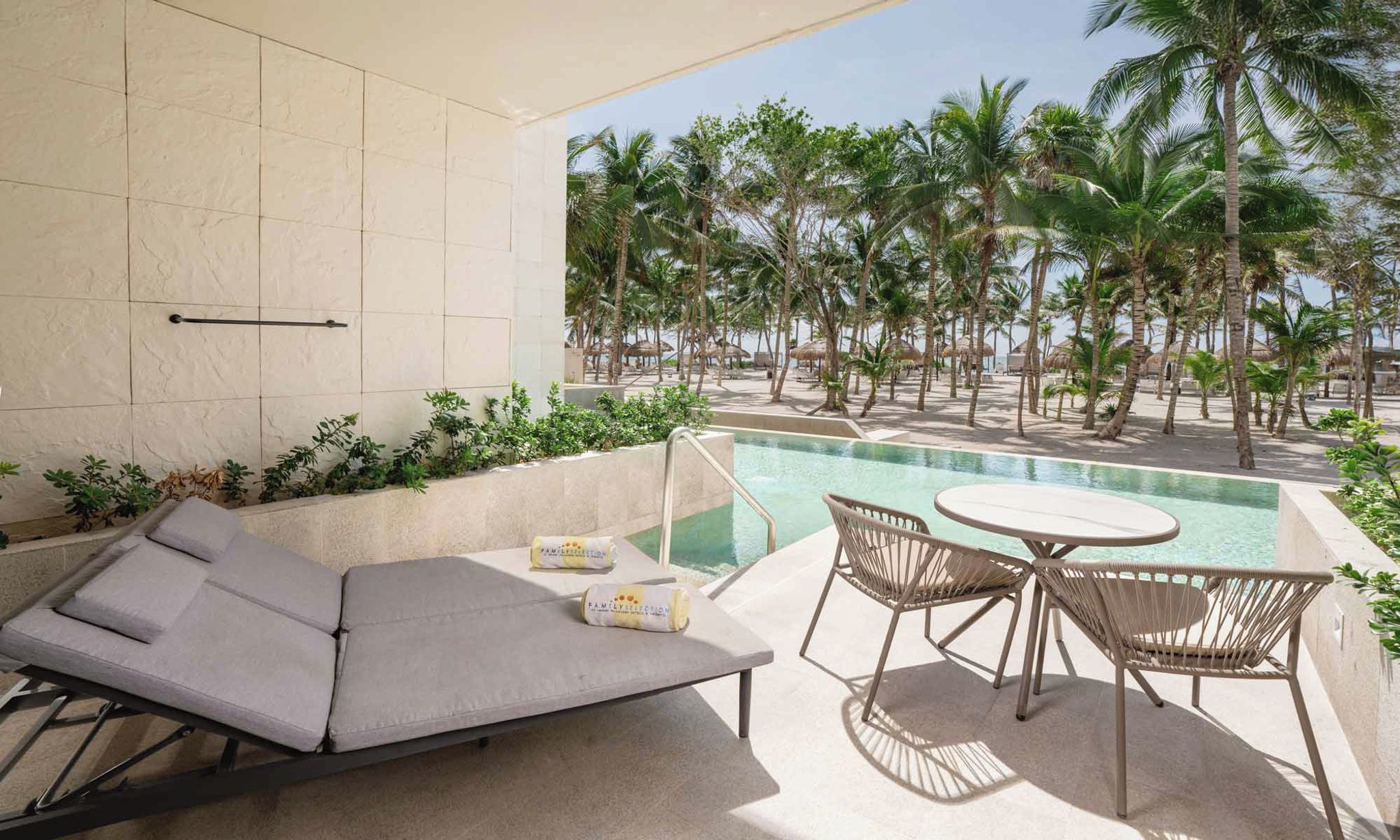 A poolside area with a lounge chair, a table, and chairs, overlooking a beach with palm trees.