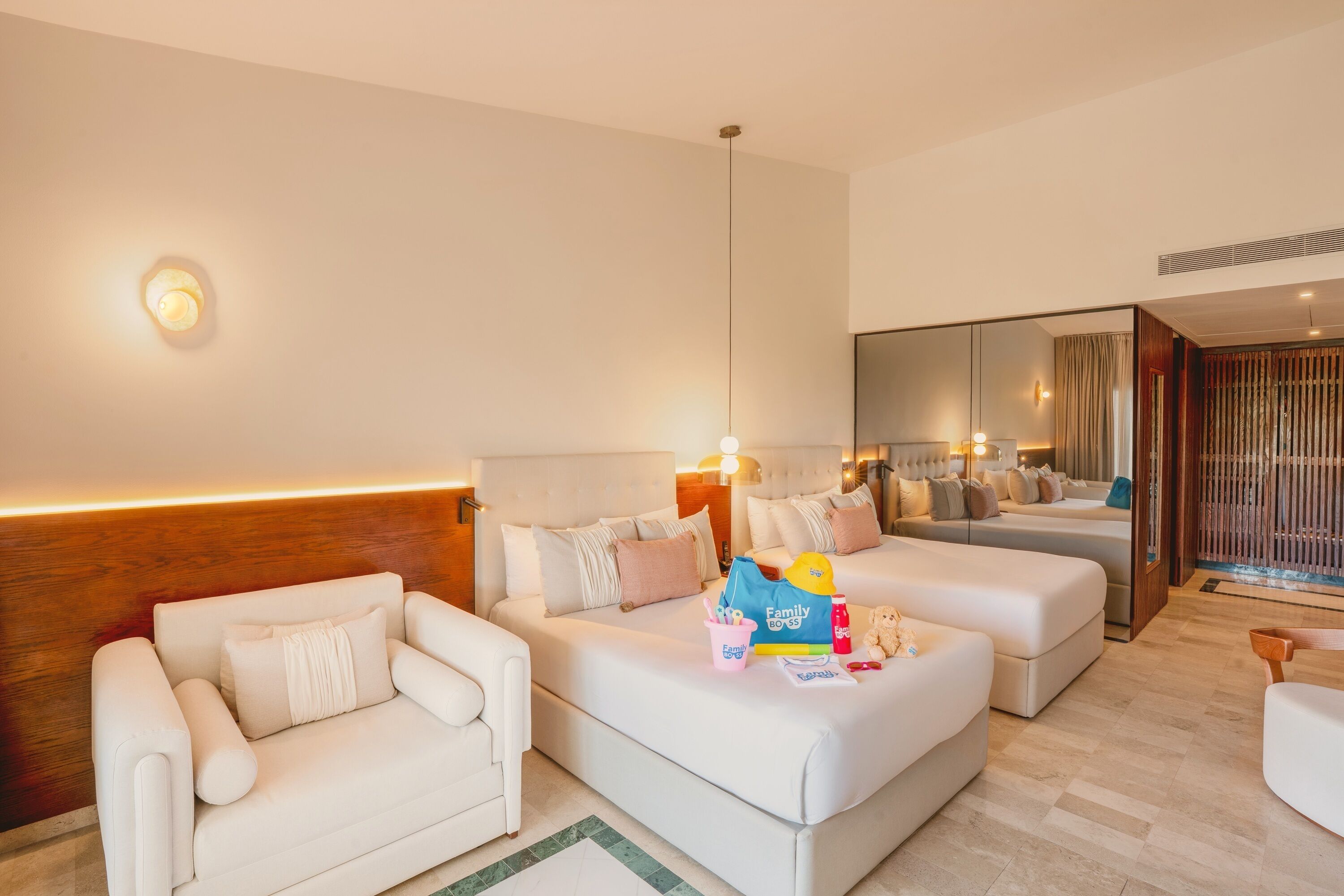 A modern hotel room with a sofa, a chair, a small table, and a bed. There is a mirror on the wall and a window with curtains.