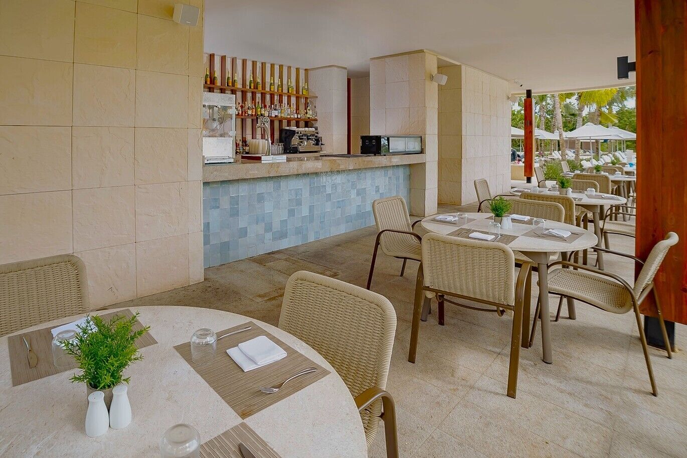 A restaurant with round tables and wicker chairs, a bar area with shelves of bottles, and a view of a pool area.
