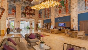 A spacious lobby with a high ceiling, a central seating area with sofas and chairs, a reception desk, and a large mural on the walls.
