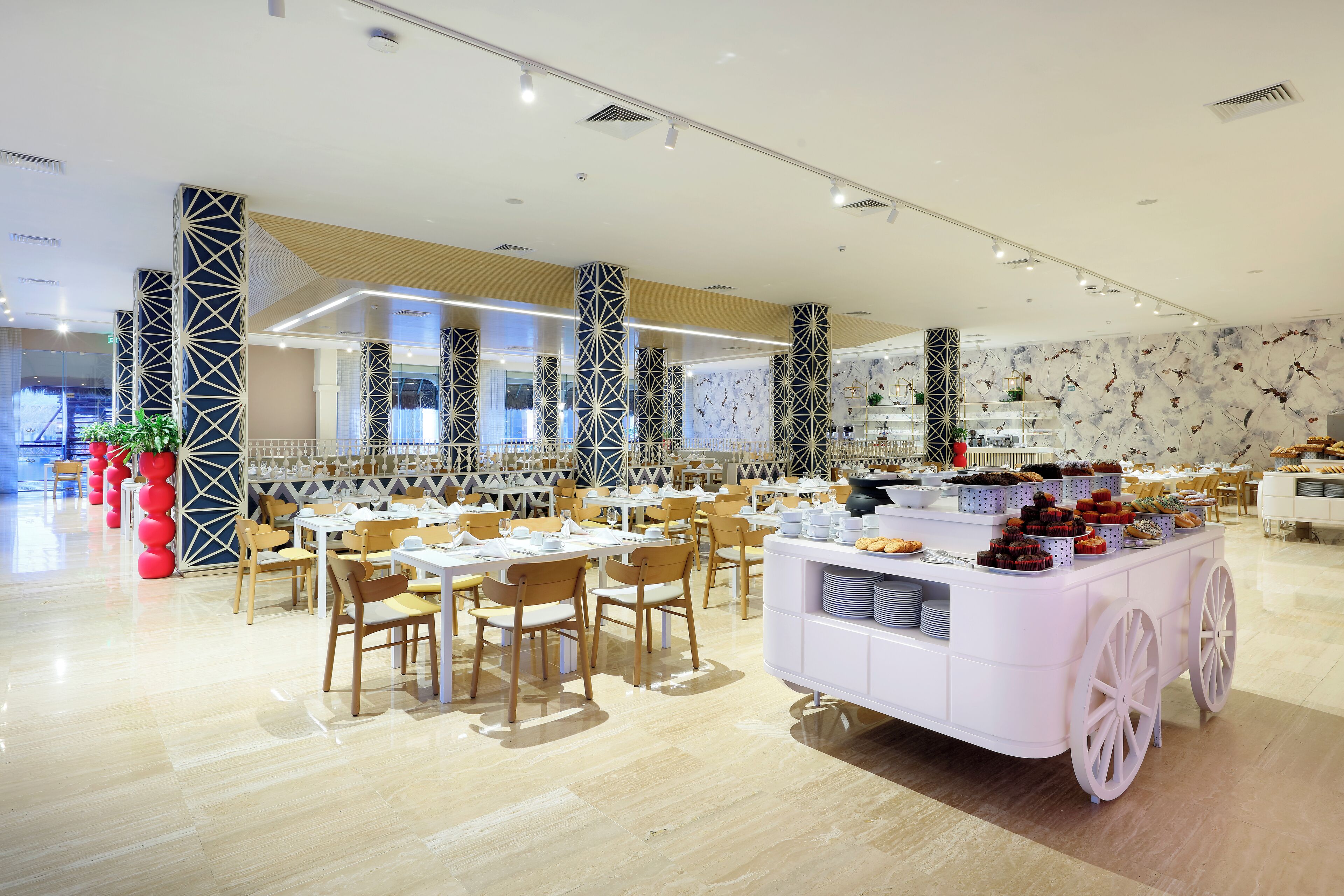 Un comedor moderno con mesas, sillas, un carrito de exhibición de alimentos y columnas decorativas.