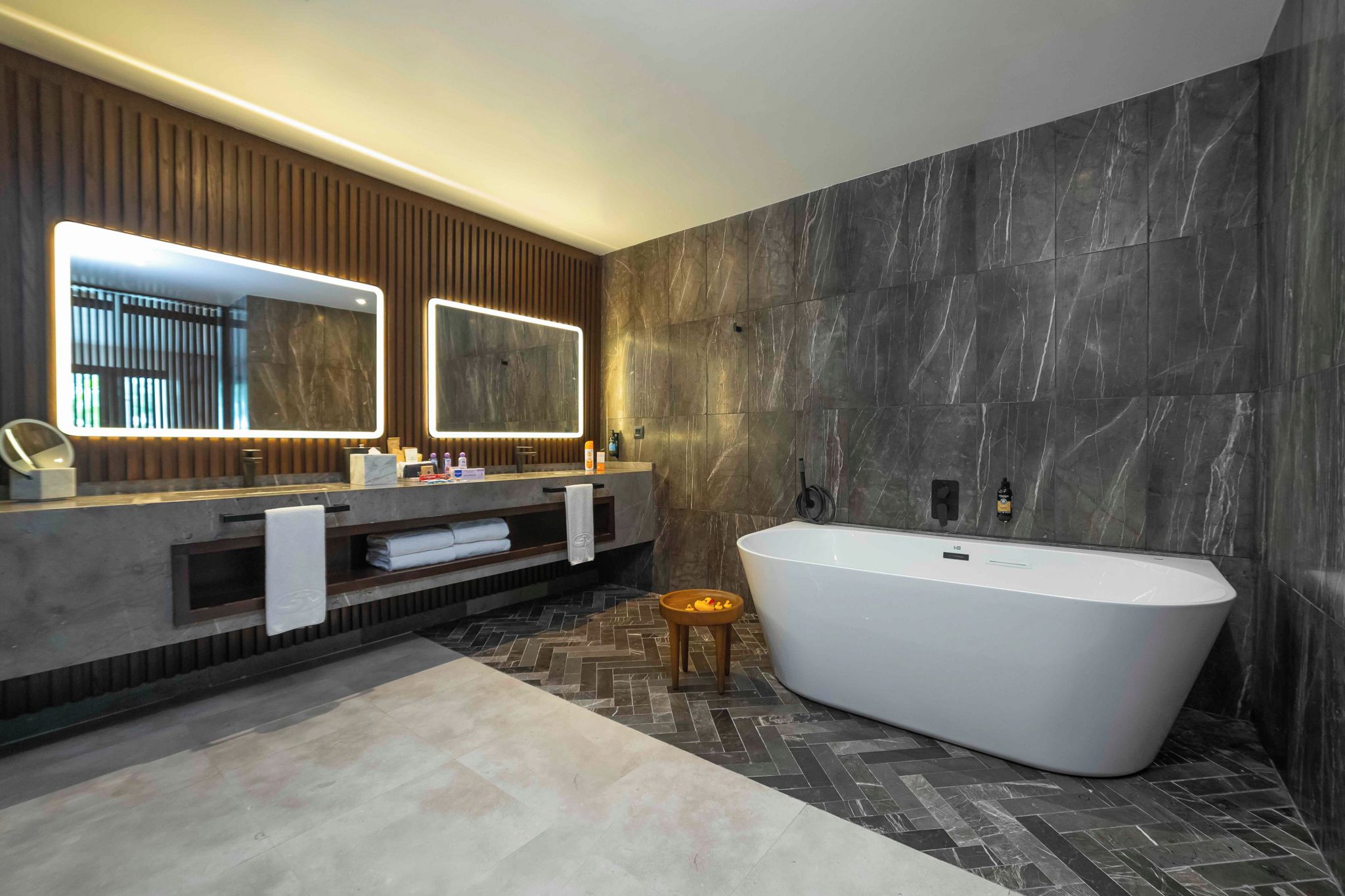 A modern bathroom with a freestanding bathtub, dual vanity mirrors, and a herringbone-patterned floor.