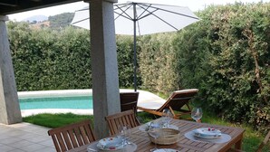 Et træbord dækket til fire personer med tallerkner, glas og en kurv, under en overdækket terrasse med en pool og grøn bevoksning i baggrunden.