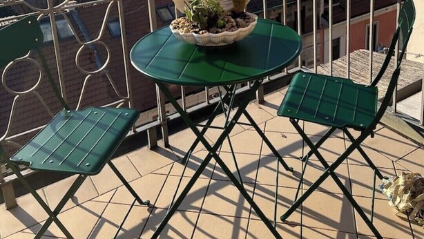 A balcony with a round table and two chairs, a potted plant, and a view of surrounding buildings.