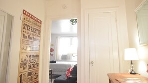 A hallway with a poster advertising a show, a door leading to a room with a bed and a window, and a lamp on a table. - 6647 2W | Enchanting Studio in St. Louis (St. Louis)