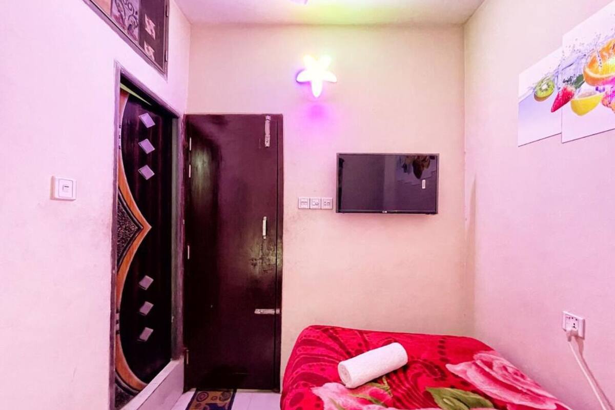 Ein kleiner, schmaler Raum mit Bett, einer Tür, einem an der Wand befestigten Fernseher und einer Deckenleuchte.